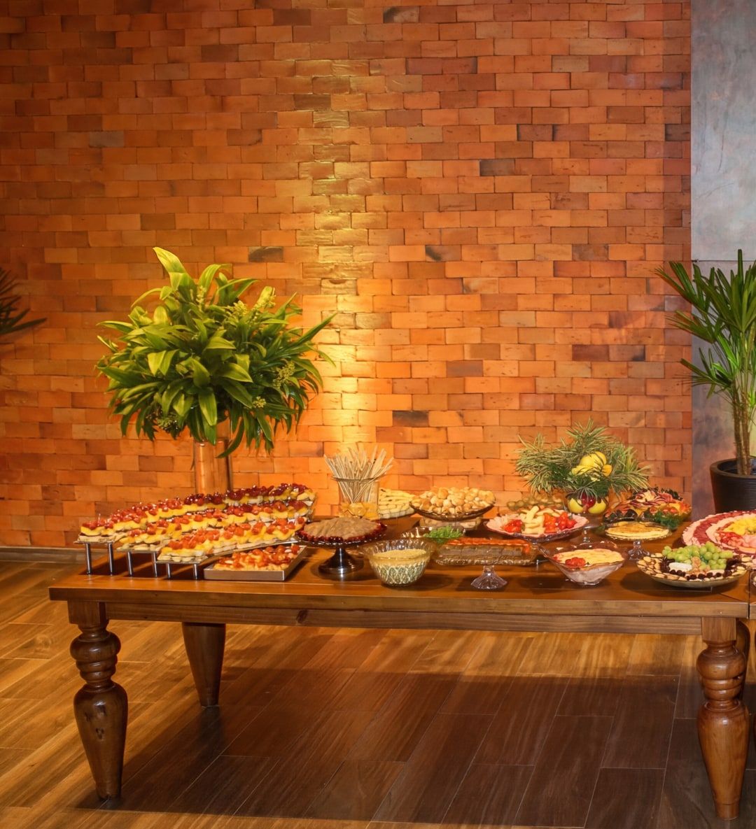 Mesa de buffet montada sobre aparador de madeira, com salgados, frios e arranjos de plantas